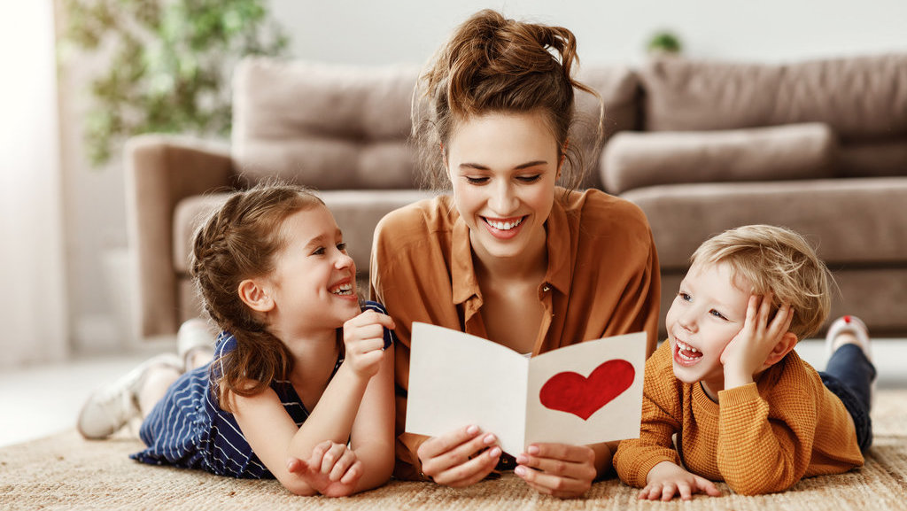 Ibu muda yang gembira dan anak kecil berbaring di lantai dekat sofa dan berpelukan sambil membaca keinginan dan selamat dengan liburan di kartu pos yang disajikan di rumah. (FOTO/iStockphoto) Hari Ibu