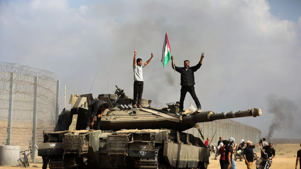 Warga Palestina mengibarkan bendera nasional mereka dan merayakannya dengan tank Israel yang hancur di pagar Jalur Gaza selatan timur Khan Younis pada Sabtu, (7/10/2023). (Foto AP/Yousef Masoud) 100 Hari Genosida Palestina