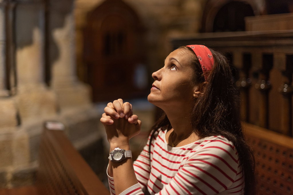 Side view of serious Hispanic female with long dark hair giving prayer with clasped hands while sitting on bench in old Catholic church and looking up Doa Kerahiman Ilahi